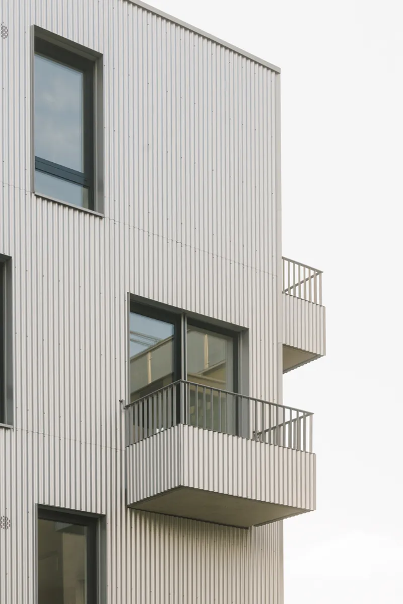 White corrugated metal facade with staggered black-framed balconies and windows under an overcast sky