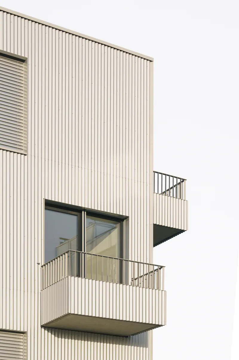Close-up of corrugated facade showing sliding glass doors opening onto cantilevered balconies with metal railings