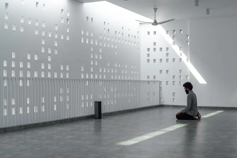 Interior wall with grid of square openings and vertical slat panel casting angular sunlight across the floor