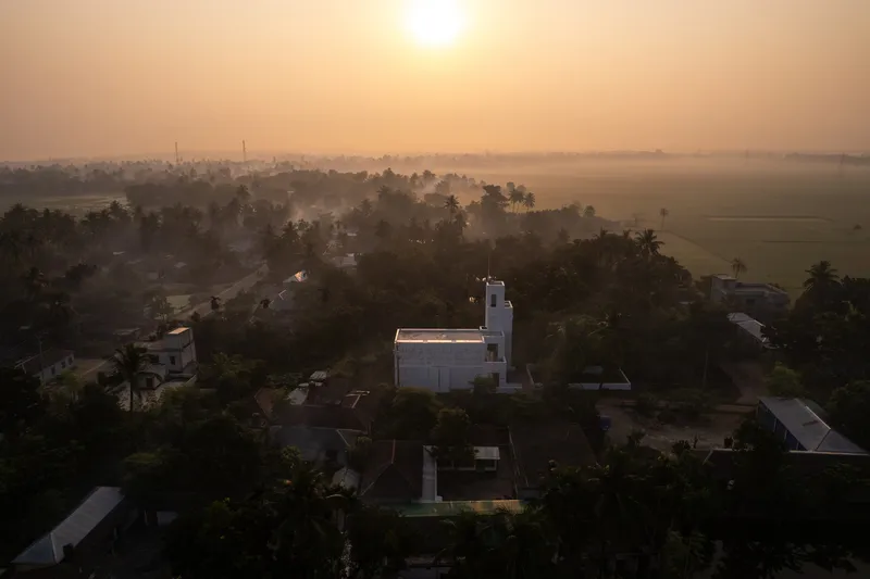 Aerial view of a white tower emerging from dense palms and houses at sunrise with morning mist