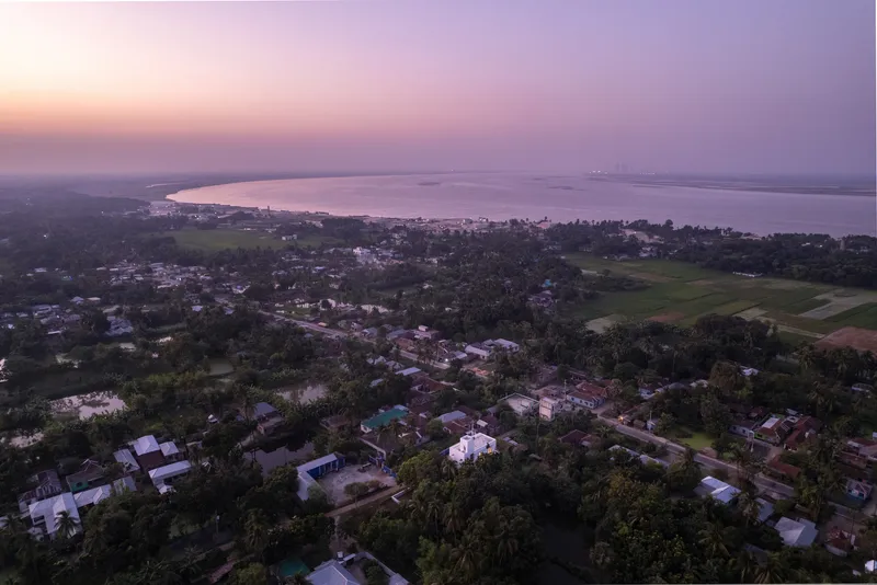 Drone view of scattered houses amid tree cover and rice fields at dusk near a waterway