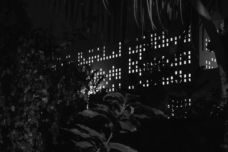 Night view of the pierced wall glowing through surrounding garden foliage