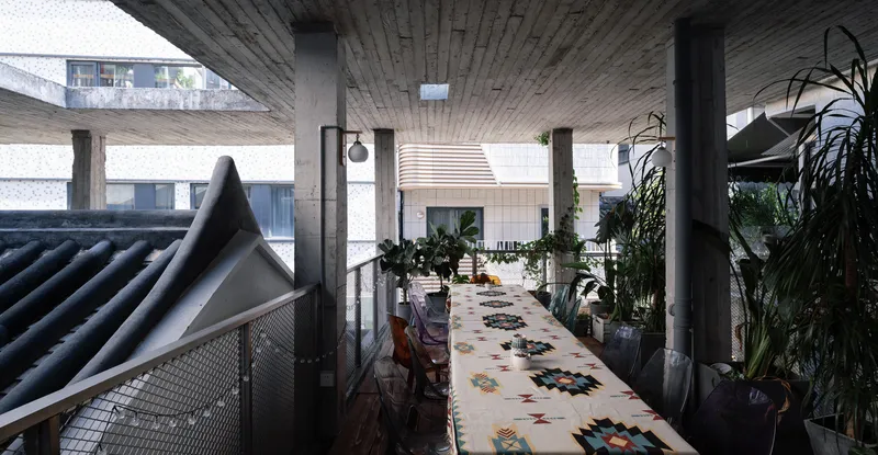 Covered corridor with patterned runner rug, potted plants, and board-formed concrete ceiling with rectangular openings overhead