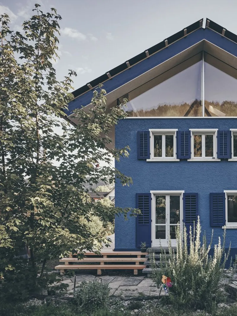 Garden facade with blue plaster walls, white-framed windows, dark shutters and timber bench beneath a young tree