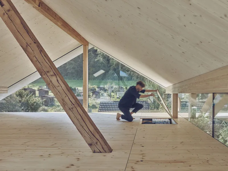 Attic room with plywood flooring, sloped timber beam, triangular window and figure working on the floor