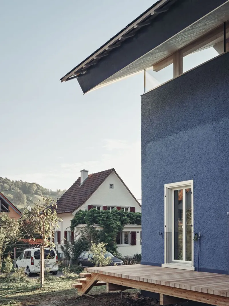 Blue rendered facade with timber deck and white-framed door overlooking neighboring gabled house