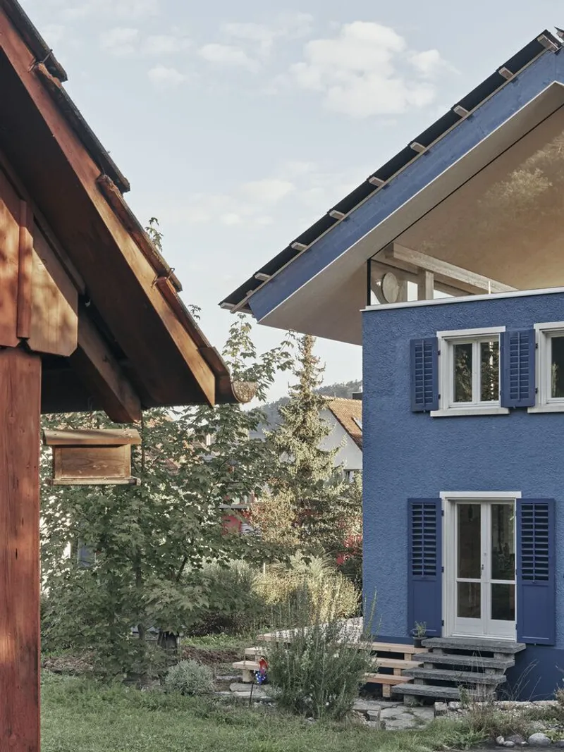 Blue stucco facade with white shuttered windows beside planted garden and timber structure