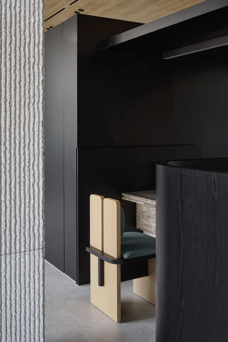 Detail of light wood chair with upholstered seat next to textured concrete column and dark millwork