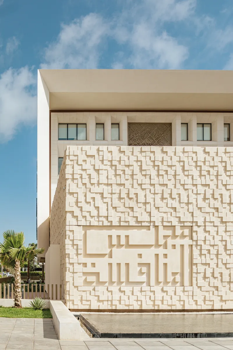 Textured stone wall with basketweave pattern and Arabic calligraphy beside a reflecting pool and palms