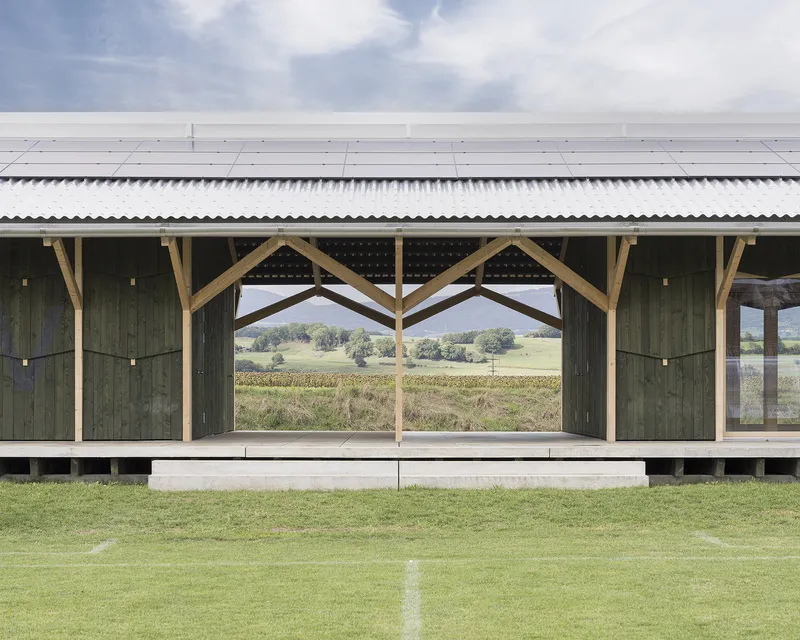 Open pavilion structure with timber A-frame columns framing a view across a playing field to distant hills