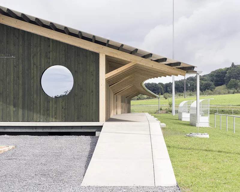 Covered walkway with exposed timber rafters and circular window opening beside a sports field