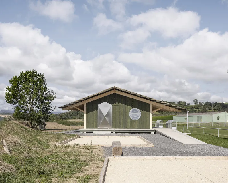 Green timber-clad gable end with diamond and circular openings on a gravel path under cloudy skies