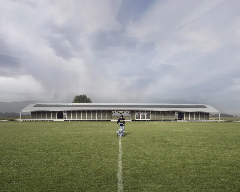 Long pavilion structure with corrugated metal roof viewed from the pitch with a figure walking toward it