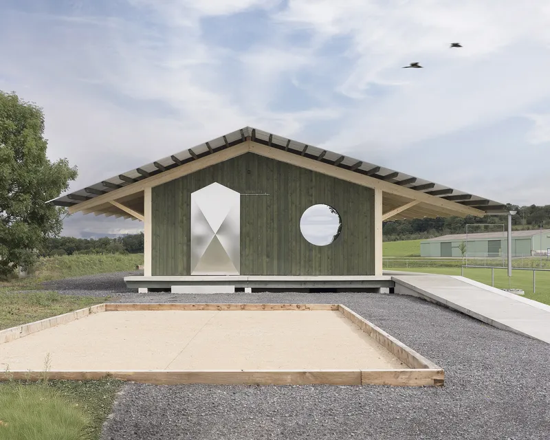 Gable facade with diamond and circular windows beside a sand pit with two birds flying overhead
