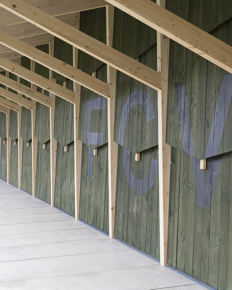 Covered walkway showing numbered green timber doors framed by exposed rafters and clerestory glazing