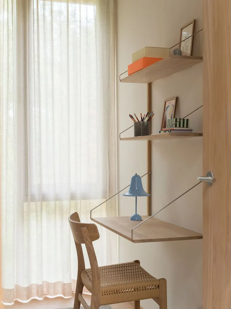 Wall-mounted desk shelves with wire rail brackets and cane chair beside a draped window