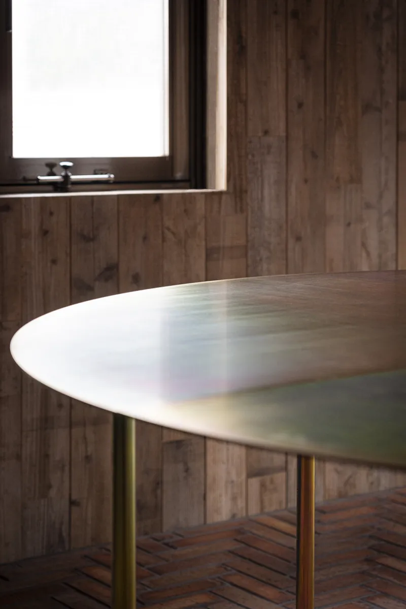Oval marble tabletop on brass legs against vertical timber cladding with diffused daylight from a window
