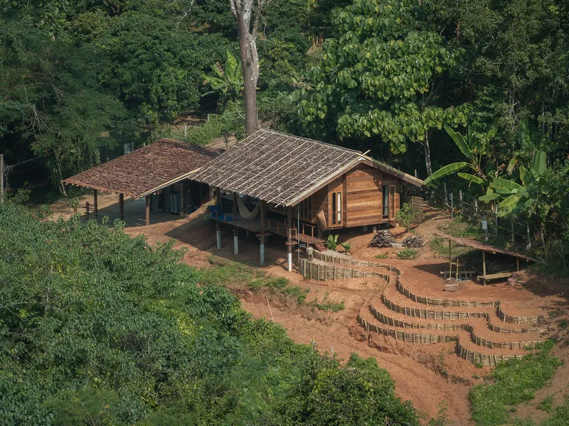 Aerial view of timber pavilions with thatched roofs and terraced bamboo fencing on red earth among tropical trees