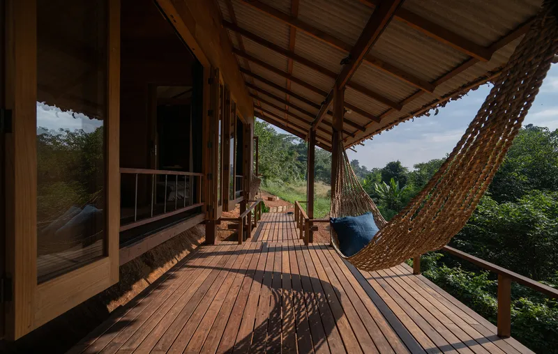 Covered timber deck with woven hammock and exposed rafters overlooking forested hillside under cloudy sky