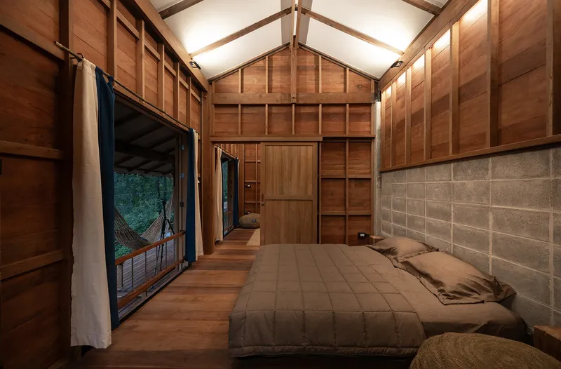 Bedroom with timber-paneled walls, exposed truss ceiling, and glazed doors opening to deck with hanging hammock