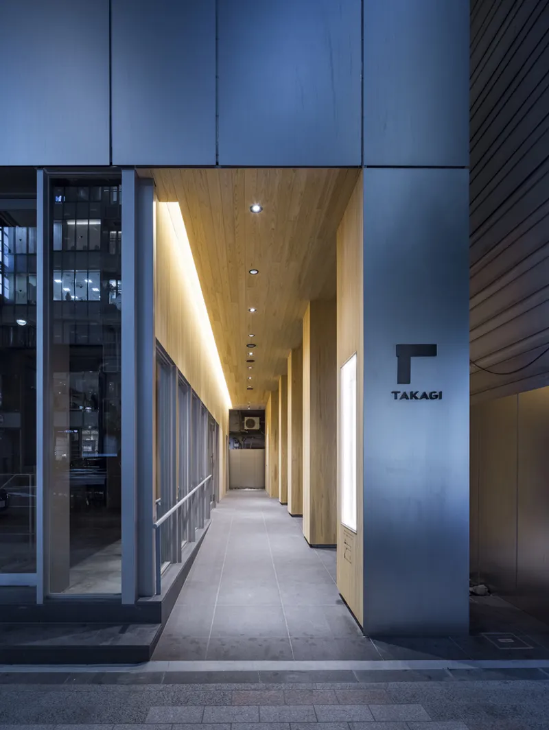 Covered entry passage with timber ceiling, backlit vertical panels, and grey tile flooring at evening