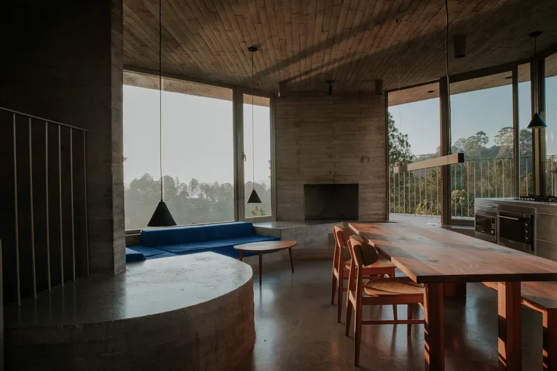 Open-plan interior showing circular fireplace, dining table, and glazed walls overlooking forested hillside in mist