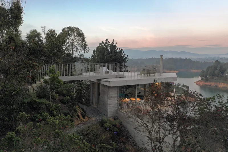 Hillside facade with cantilevered roof terrace overlooking a reservoir at dusk with layered mountain ridges beyond