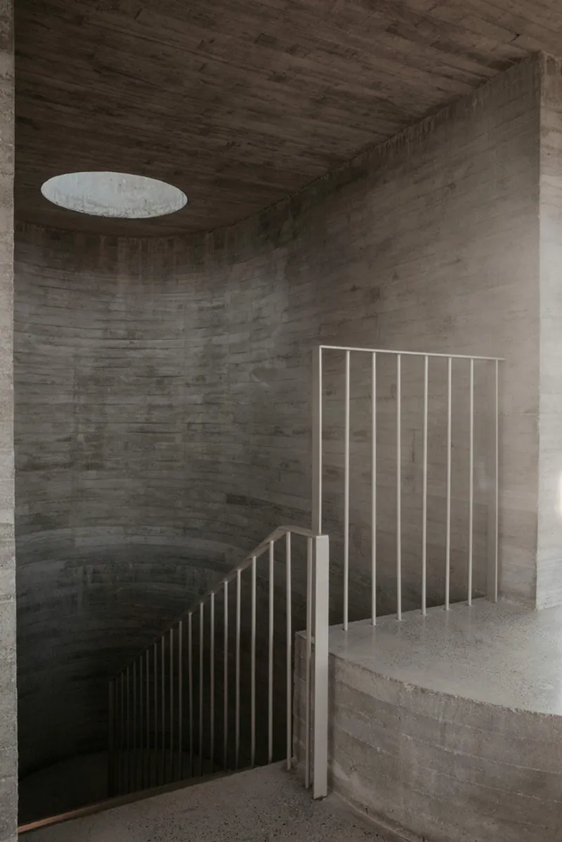 Curved concrete stairwell with vertical metal balustrade and circular skylight above board-formed walls