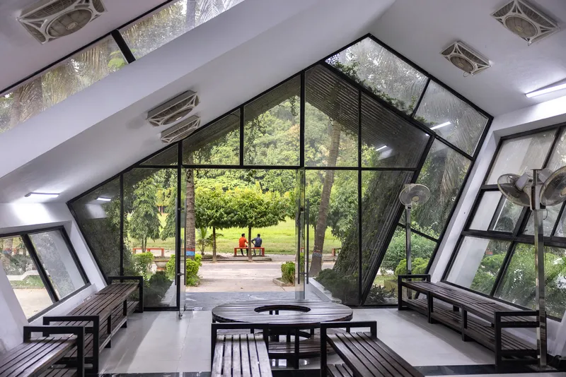 Interior view through the angled glass wall framing wooden benches and park beyond