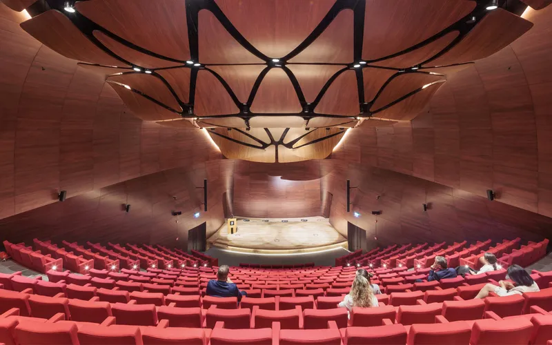 Concert hall interior with red seating rows facing a wood-paneled stage under branching structural beams