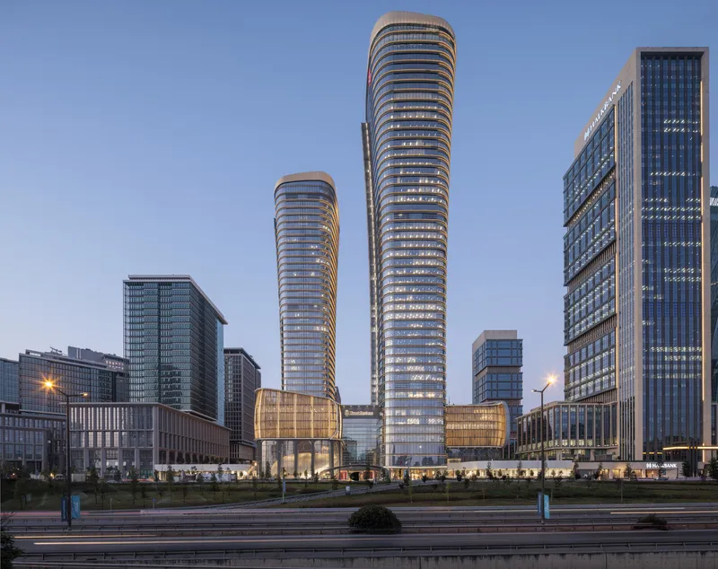 Two cylindrical glass towers with horizontal banding rise above a podium base at dusk