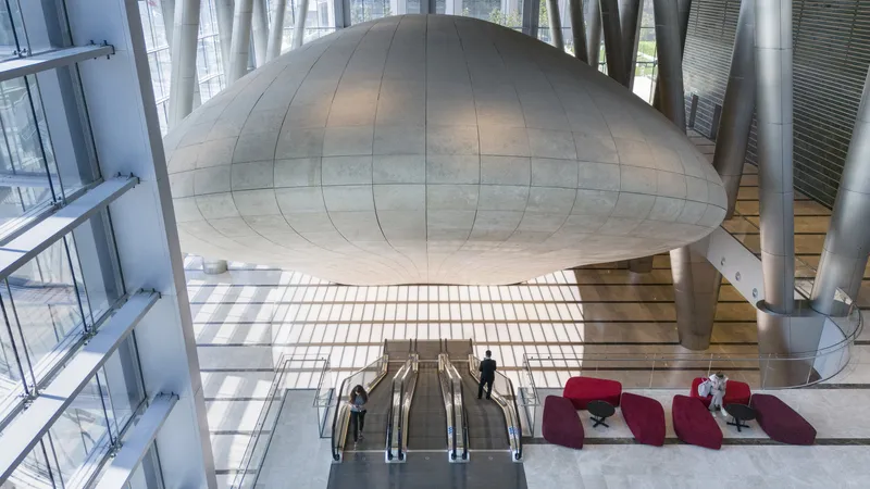 Double-height lobby with suspended dome-shaped volume and escalators beneath a slatted glass ceiling