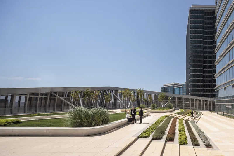 Landscaped plaza with terraced planted beds and visitors walking toward a horizontal louver facade