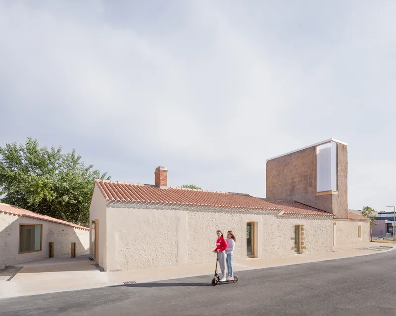 Low stone building with terracotta tile roof and vertical concrete tower as two figures stand in the street