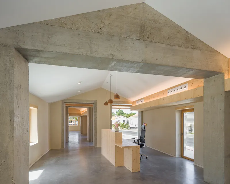 Interior space with exposed concrete beam framing timber desk and pendant lights under vaulted ceiling
