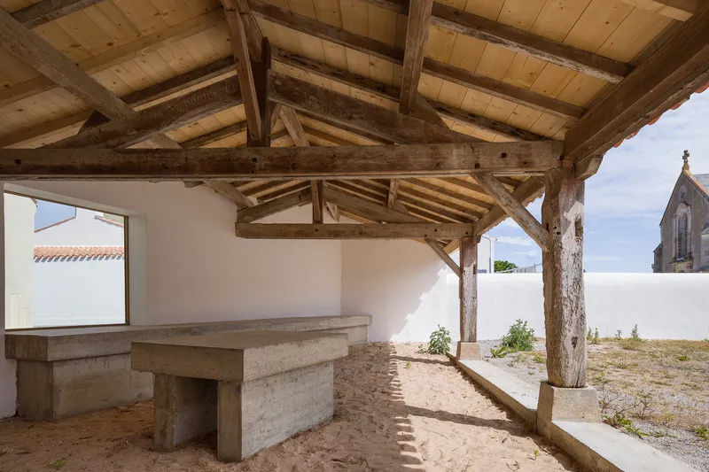 Covered outdoor terrace with exposed timber trusses and stone benches set in sandy ground