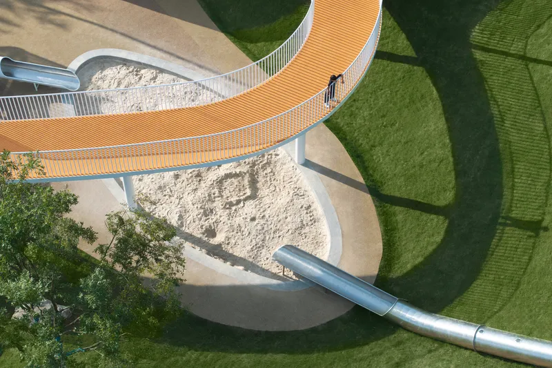 Aerial view of curved orange bridge connecting sand play areas with metal slide and lawn below