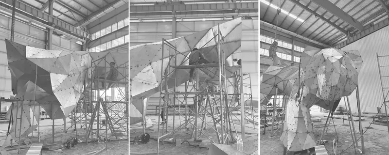 Faceted metal installation suspended on steel scaffolding inside an industrial fabrication warehouse