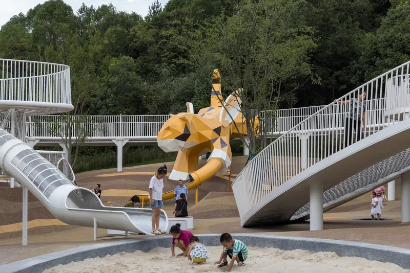 Faceted yellow sculpture rising from sand play area beneath converging white walkways and forest backdrop