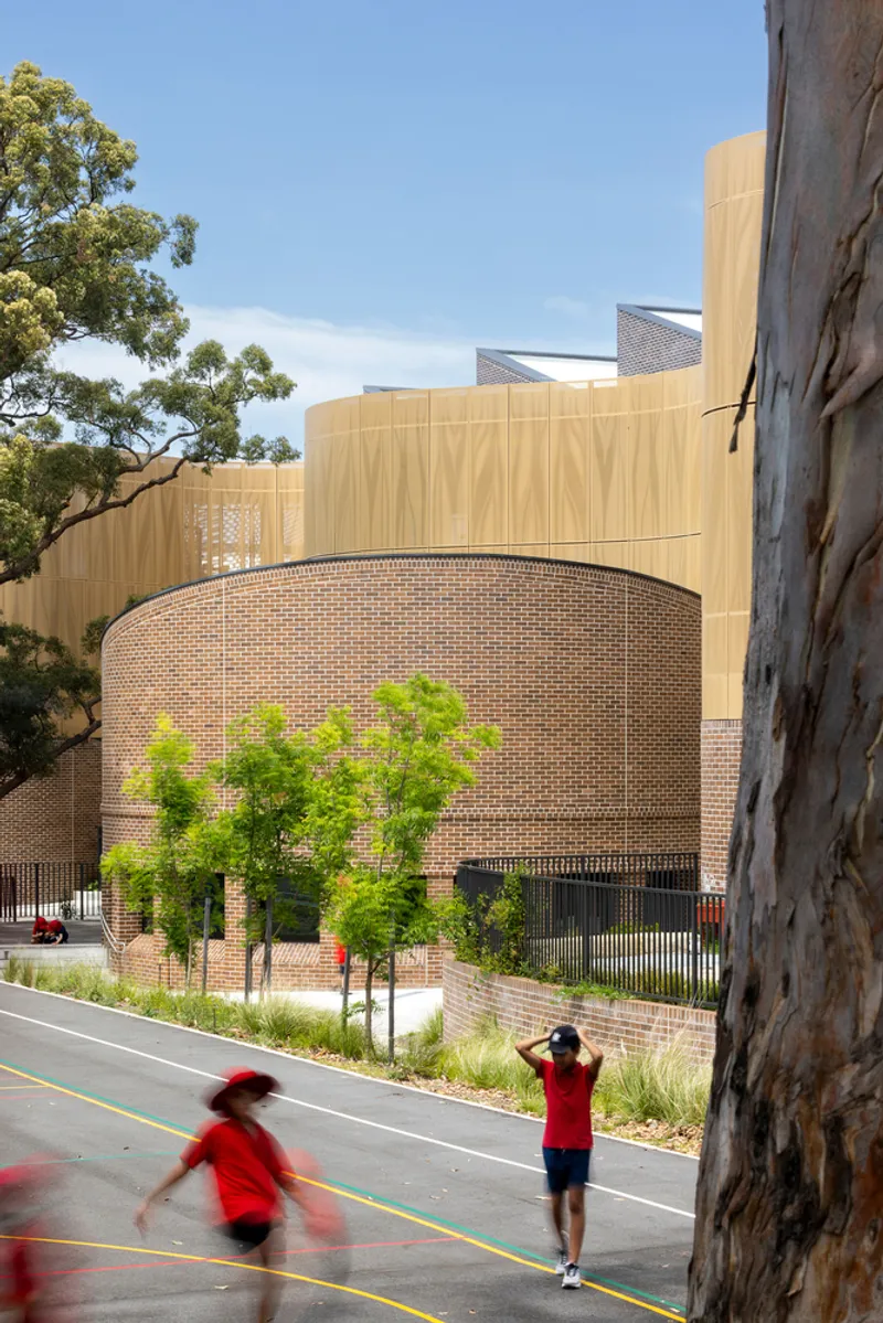 Street view of curved brick wall with planted buffer and timber-clad upper volume beside eucalyptus trunks