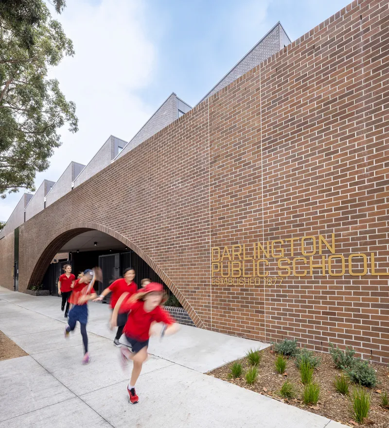 Curved brick facade with arched entry and metal signage as students in red uniforms run along the path