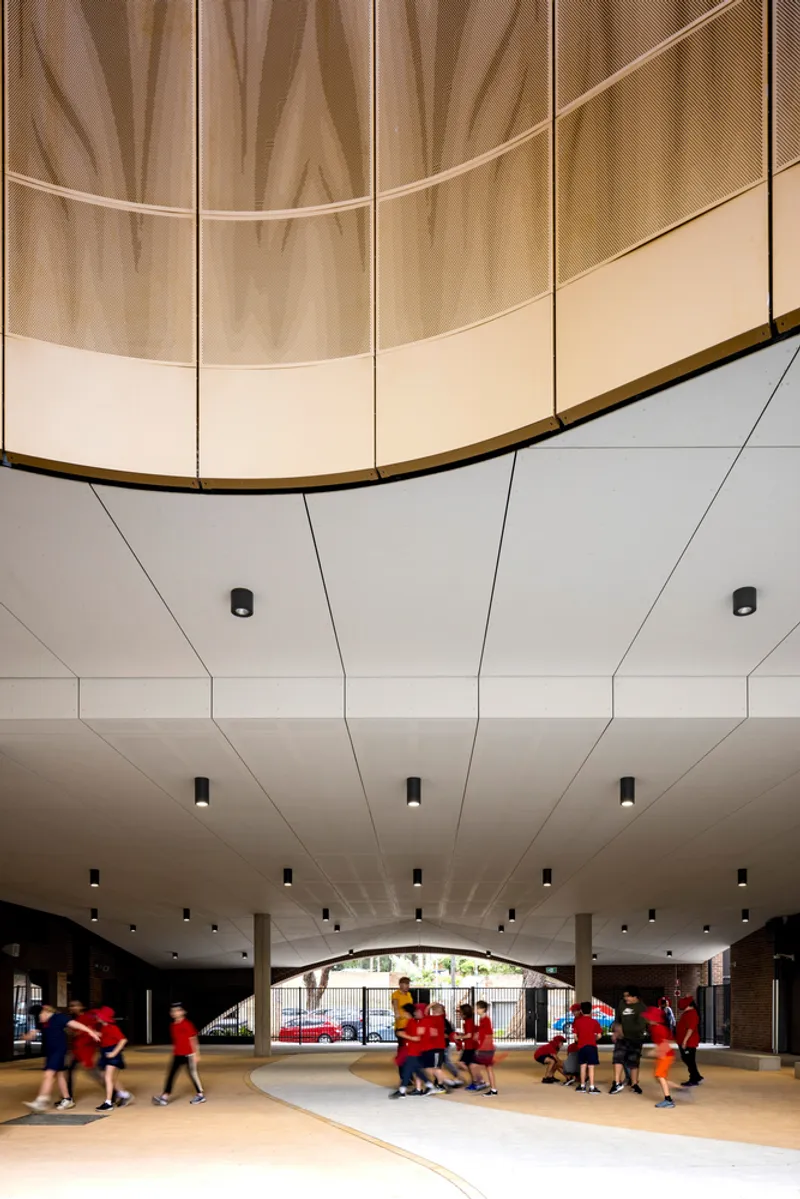 Interior view of curved perforated metal and timber screen suspended above white paneled ceiling with students below