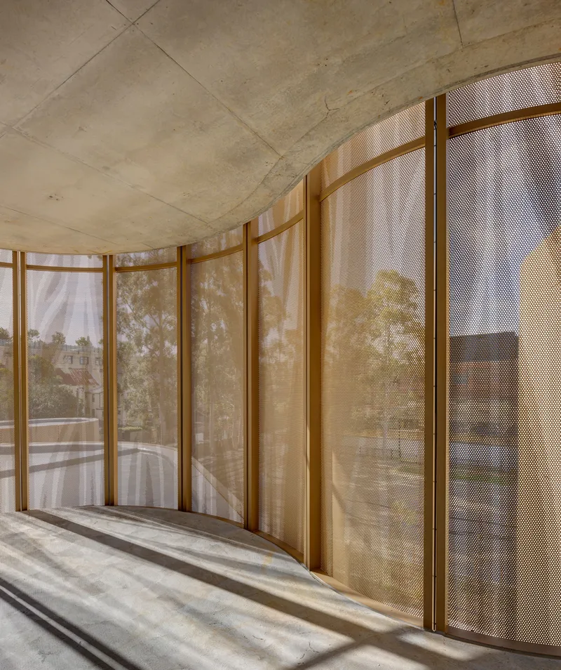 Curved perforated metal screen wall with timber framing filtering dappled sunlight onto concrete floor