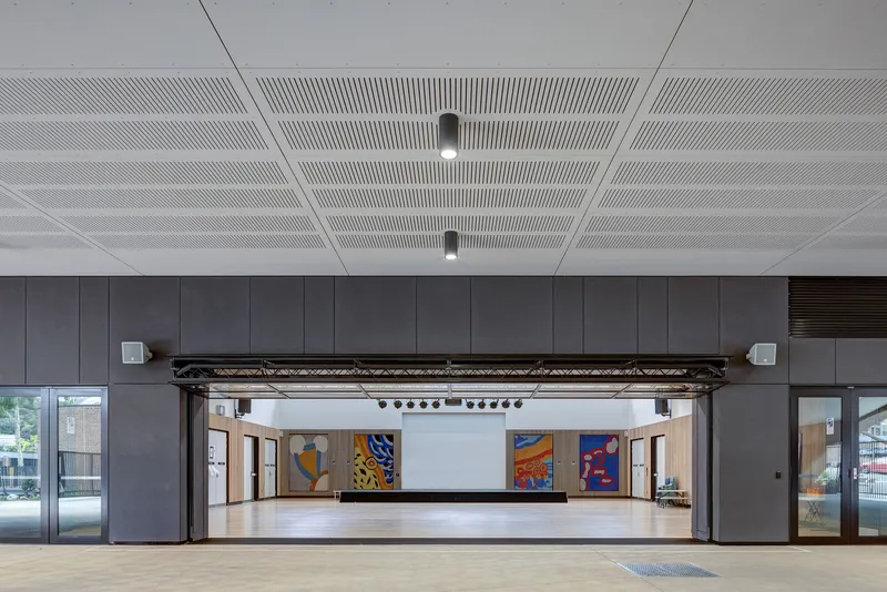 Interior hall with perforated white ceiling showing raised stage flanked by grey paneled walls and track lighting