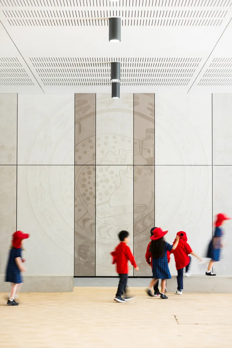 Plywood wall panels with carved indigenous motifs beneath a perforated white ceiling as children walk past
