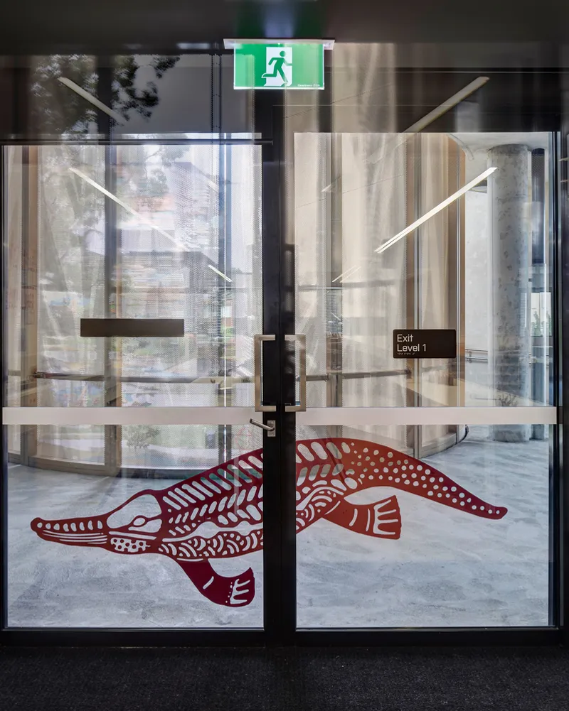 Glass exit door with indigenous animal decal in red and exit signage reflecting courtyard beyond