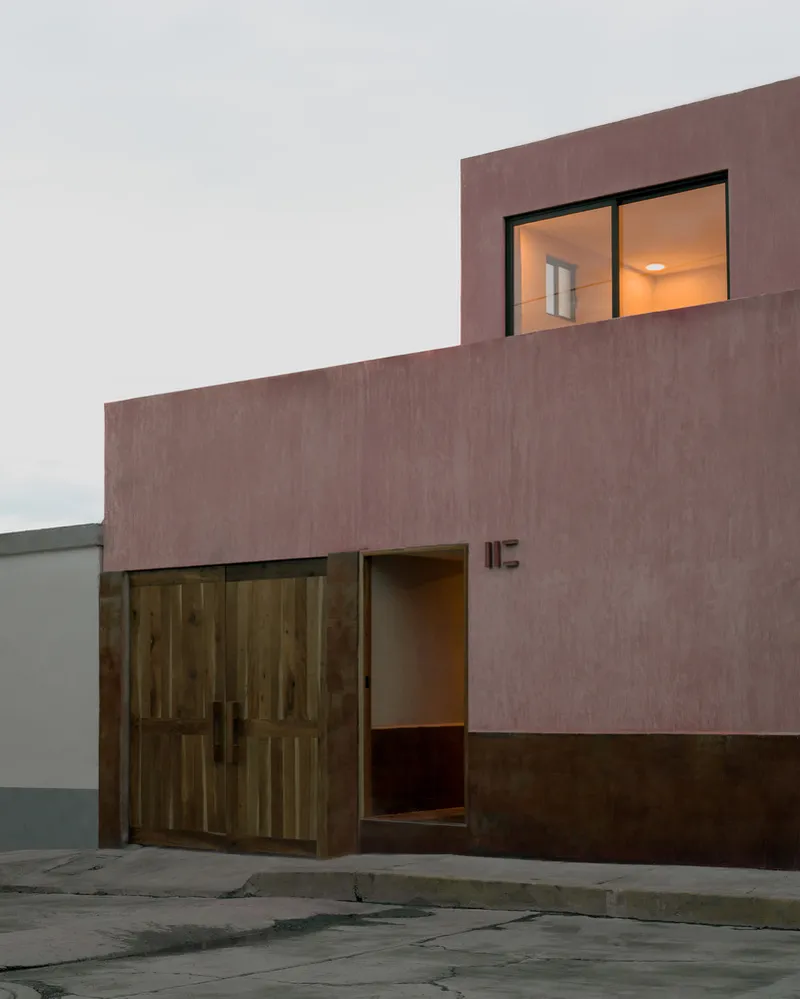 Street facade in textured pink render with timber door and illuminated upper window at dusk
