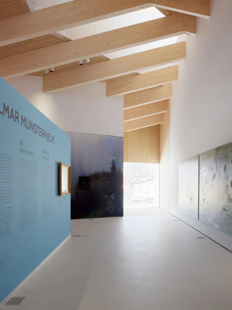 Gallery corridor with exposed timber beams and skylights illuminating a blue exhibition wall and displayed artworks