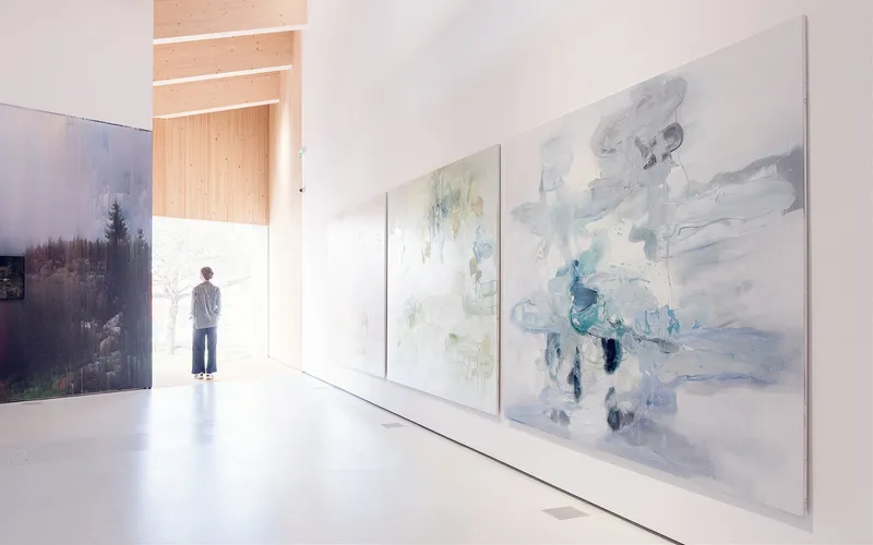 Gallery interior with visitor standing in backlit timber portal looking out to daylight beyond painted canvases