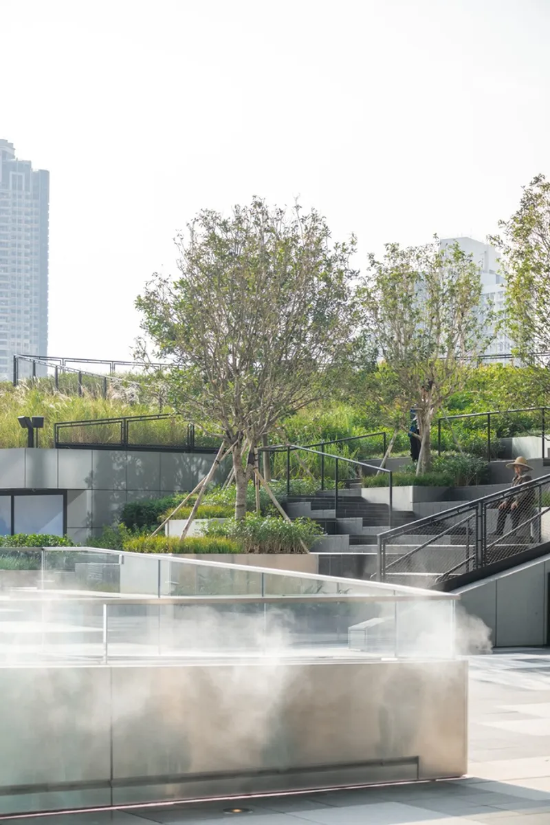 Tiered outdoor courtyard with steel planters emitting mist beneath trees and metal railings